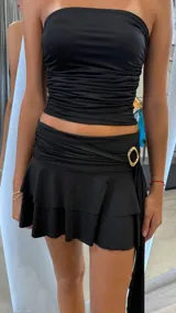 Minifalda negra con efecto drapeado en la cintura y detalle de hebilla dorada en forma de anillo. La falda tiene un ruedo con volados.