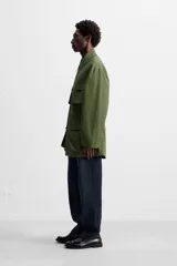 Parka verde militar de tejido técnico ligero, corte relaxed fit, cuello solapa y manga larga. Tiene cuatro bolsillos de plastrón con solapa en el delantero, cintura ajustable en el interior y cierre frontal con cremallera oculta por solapa con botones a presión.