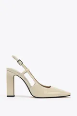 Zapato de tacón destalonado color beige acharolado, con punta fina, tacón ancho en bloque y cierre mediante tira trasera con hebilla ajustable.
