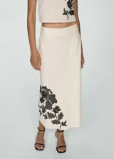 Falda midi color crema, de tiro alto y corte recto. Presenta bordado de flores negras en el lateral inferior.