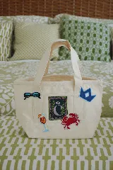 Bolso tote pequeño de lona color crudo con doble asa corta. Decorado con parches termoadhesivos de gafas de sol, inicial con corona, cóctel y cangrejo.