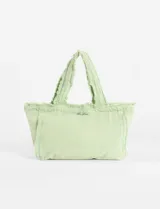 Bolso tipo tote de lona color verde claro, con efecto lavado o desgastado y bordes desflecados en las asas y la abertura superior. Presenta un bordado pequeño en el centro frontal.