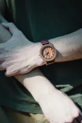 Reloj de pulsera con caja, esfera y malla de madera de sándalo rojo, con marcadores y manecillas negras. Presenta un diseño circular con corona lateral.