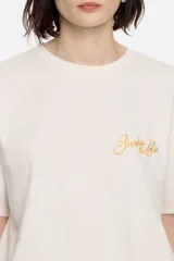 Camiseta color crudo de algodón, con cuello redondo y mangas cortas. Presenta logo bordado en color dorado en el frente.