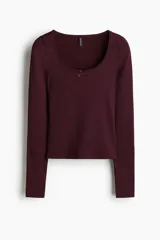 Remera de manga larga ajustada al cuerpo, confeccionada en tejido acanalado color bordó. Presenta un escote pronunciado ribeteado en encaje y un pequeño moño de satén en el centro del escote.