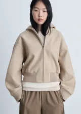 Chaqueta beige con capucha, cierre frontal con cremallera, puños y cintura elásticos.