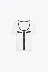 Camiseta blanca de corte slim fit, con cuello redondo y mangas cortas. Presenta detalles en negro en el cuello, los bordes de los falsos bolsillos y la botonadura frontal con botones dorados.
