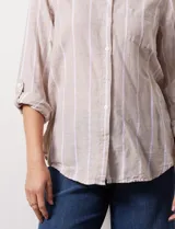 Camisa color beige con rayas verticales blancas, de lino, con cuello con solapa y abertura central con botones. Tiene un bolsillo aplicado en el pecho y mangas largas regulables con presilla y botón.