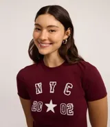Remera corta color bordó de algodón con elastano, cuello redondo y mangas cortas. Estampa frontal con letras "NYC" y el número "2002" con una estrella en el medio.