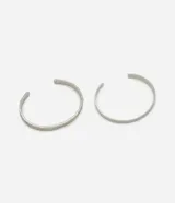 Kit de dos brazaletes rígidos de metal reciclado con baño de níquel. El conjunto incluye un brazalete de superficie lisa y minimalista, y otro con textura martillada. Ambos cuentan con una abertura posterior para facilitar su colocación y ajuste.