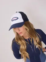Gorra de lona blanca con visera azul marino y malla trasera azul marino. Presenta un logo bordado en el frente con letras rosadas y contorno azul marino.