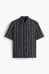Camisa de manga corta con rayas verticales blancas sobre fondo negro, cuello inglés, tapeta con botones y bolsillo de parche en el pecho. Corte recto y holgado.