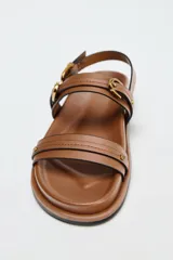 Sandalias planas color marrón con doble tira delantera con detalle de hebilla y cierre mediante hebilla en la pulsera al tobillo.