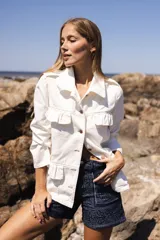 Chaqueta de denim liviana en color beige con detalle de volados en el frente, cuello camisero y bolsillos funcionales. Presenta un calce relajado y cierre frontal mediante botones.