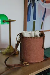 Cartera tipo canasta color terracota con textura símil piel de cocodrilo, con forro de tela blanca ajustable con cordón. Tiene correa larga de cuero marrón con hebilla dorada.