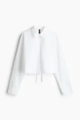 Camisa cropped blanca de popelina de algodón, con cuello, botones, canesú en la espalda, mangas largas con tapeta y puños abotonados. Abertura pronunciada con tiras doble en la espalda.