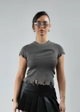 Remera gris de tejido de punto acanalado con cuello redondo, mangas cortas con borde desflecado, logo en el frente y ruedo irregular con terminación desflecada.