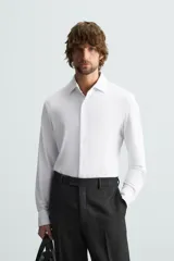 Camisa color beige de corte slim fit, confeccionada en tejido elástico que minimiza las arrugas. Tiene cuello italiano abierto, manga larga con puños abotonados y cierre frontal con botones.