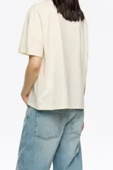 Camiseta oversize de algodón y poliéster color crema con escote redondo y mangas cortas anchas. Presenta un bordado frontal con hilos colgantes de colores variados.