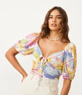 Top cropped de tejido tipo laise con estampado floral pixelado en tonos pastel (rosa, amarillo, azul, blanco). Presenta escote corazón con abertura central y lazo ajustable, y mangas cortas abullonadas.
