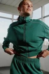 Campera deportiva verde esmeralda con cierre frontal y cuello alto. Presenta detalles de ribete blanco en hombros y mangas.