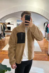 Campera marrón de tela tipo borreguito con cierre frontal y bolsillo de parche negro en el pecho.
