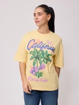Remera color crudo de corte oversized con estampado de palmeras, flores y texto 'California' y 'Rusty Tribe'.
