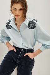 Camisa blanca de algodón con cuello clásico, mangas largas abullonadas con puños anchos y cierre frontal de botones. Presenta aplicaciones bordadas en dorado con detalles de animal print en los hombros.