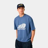 Camiseta de manga corta, corte boxy (holgado), color azul, con un estampado frontal grande en blanco que simula ondas o viento y el texto "EMESTUDIOS®" debajo.