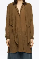 Camisa larga de corte recto, color marrón, confeccionada en tejido plisado. Presenta cuello camisero, botones frontales y mangas largas con puño. Destaca por sus maxilazos colgantes en el escote.