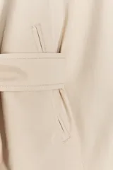 Trench corto color crema con cuello solapa, manga larga con trabilla, bolsillos de doble vivo, forro interior y cinturón lazada en el mismo tejido.