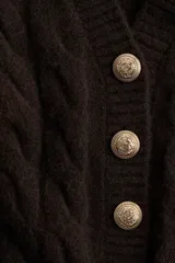 Cárdigan corto marrón oscuro en tejido de punto trenzado con lana. Tiene cuello en V, hombros ligeramente caídos, cierre frontal con botones dorados metálicos y terminaciones acanaladas en cuello, puños y bajo.