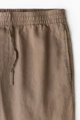 Pantalón marrón claro de lino, corte recto, con cintura elástica y cordón ajustable. Posee bolsillos laterales y trasero.