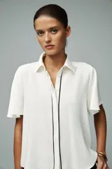 Camisa blanca de corte recto, con cuello solapa y mangas cortas. Presenta vivos negros a contraste en el frente y aberturas laterales en el ruedo. Cierre frontal con botones ocultos.