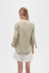 Blusa color beige con estampado de arabescos blancos, cuello mao, mangas 3/4 con lazos en los puños, cierre parcial con botones y bolsillos frontales.
