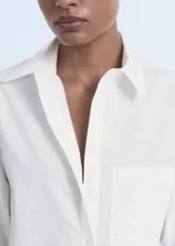 Camisa blanca de popelín de algodón, manga tres cuartos, corte crop y diseño recto. Presenta cuello camisero, bolsillo en el pecho con detalle de strass y cierre frontal con botones.