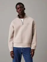 Buzo color beige de corte holgado, con cuello alto y cremallera corta. Tiene puños y bajo elásticos, y logo de Calvin Klein grabado en el pecho.