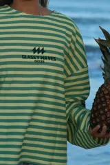 Buzo reversible de manga larga, con un lado a rayas horizontales verdes y blancas, y el otro lado liso verde. Presenta un estampado en la espalda con el texto "GLASSY WAVES" y un logo de rayos.