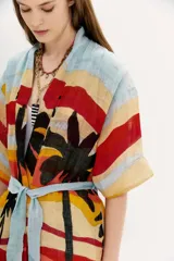 Kimono largo de lino con estampado de paisaje playero con palmeras, en tonos celeste, amarillo, rojo, verde y negro. Tiene mangas amplias, bolsillos al frente y lazo en la cintura.