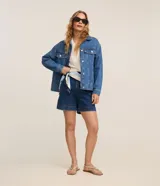 Campera trucker de jeans azul oscuro, corte amplio, con cuello clásico, cierre frontal con botones blancos, mangas largas y bolsillos delanteros con solapa.