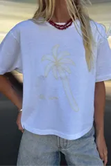 Remera blanca de algodón con cuello redondo y mangas cortas. Presenta un estampado bordado de una palmera en el centro del pecho y la inscripción "Villa Lovers" en la parte inferior.