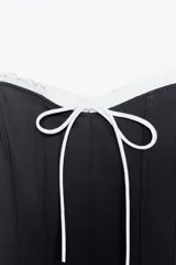 Vestido corto negro con tirantes finos ajustables, escote recto con volantes blancos y lazo decorativo. Cierre lateral con cremallera oculta.