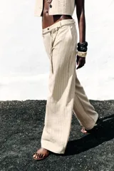 Pantalón de tiro alto color beige con finas rayas verticales negras, pernera ancha y cinturón trenzado a tono.