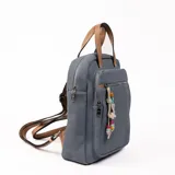 Mochila gris con detalles en marrón, con cierre superior y bolsillo frontal con cierre. Tiene manija corta superior y correas traseras regulables. Incluye llavero decorativo con cuentas de colores.