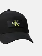 Gorra tipo trucker negra con visera curva. Presenta un panel frontal de sarga y paneles traseros de malla. En el frente lleva un parche rectangular negro con el logo de Calvin Klein en color amarillo neón.
