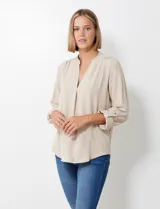 Blusa de gasa color beige, con cuello a la base y escote en V. Manga larga con presilla y botón para ajustar el largo.