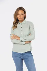 Camisa color beige de lino, de manga larga con botones metálicos personalizados y presilla en la manga. Tiene cuello clásico, cierre frontal parcial con botones y bolsillo de parche en el pecho.