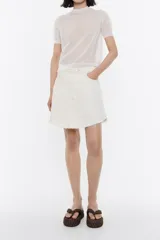 Falda mini de denim color blanco, corte línea A, cierre frontal con botón y cinco bolsillos. Tiene detalles de costuras a la vista y terminación desflecada.