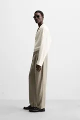 Pantalón negro de corte amplio, con pliegues frontales, bolsillos laterales y traseros de ojal. Incluye cinturón de la misma tela con hebilla metálica.