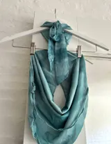 Top halter de color celeste/turquesa, con efecto drapeado y un accesorio tipo concha marina como broche central. El diseño es holgado y se ajusta en la espalda.